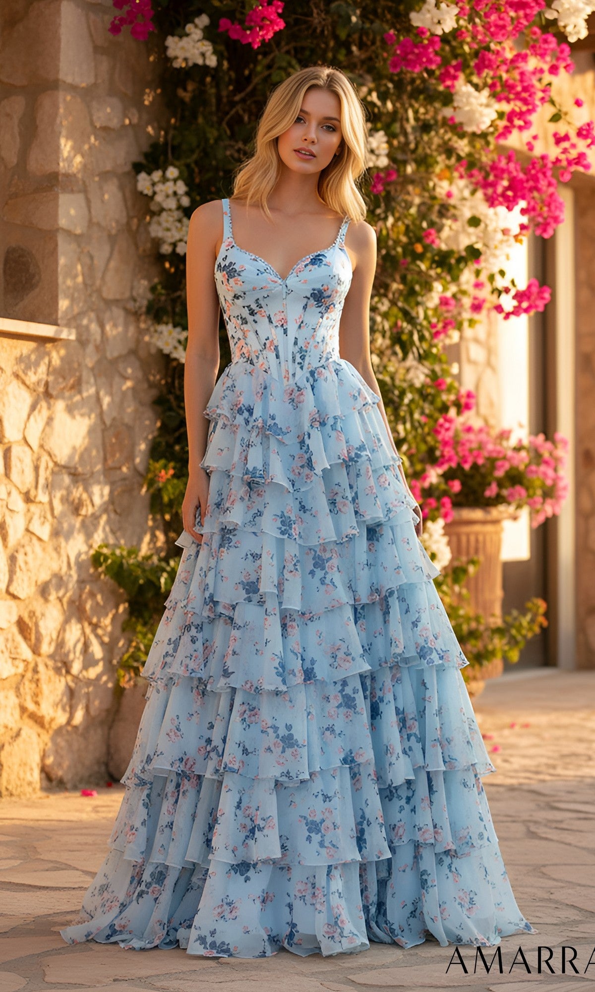 A woman wearing the Amarra 89613 long prom dress in blue/print, featuring a floral pattern and tiered ruffle chiffon skirt, stands outdoors by stone walls with blooming pink and white flowers.